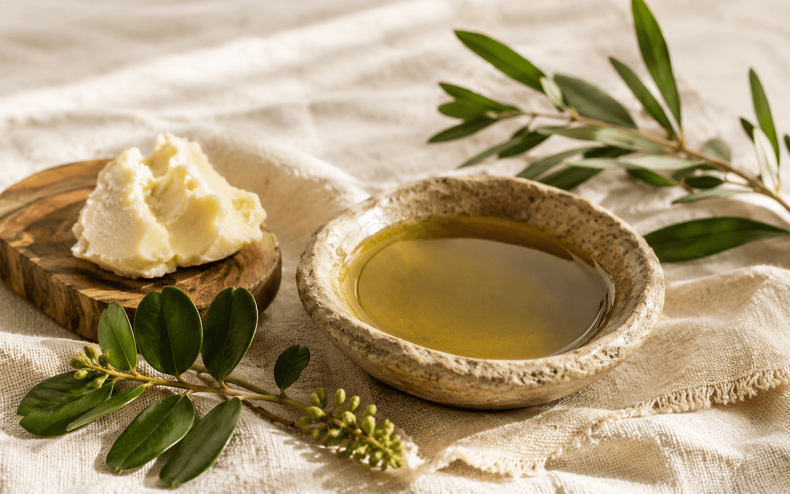 A bowl of golden oil beside shea butter and botanical sprigs on natural linen